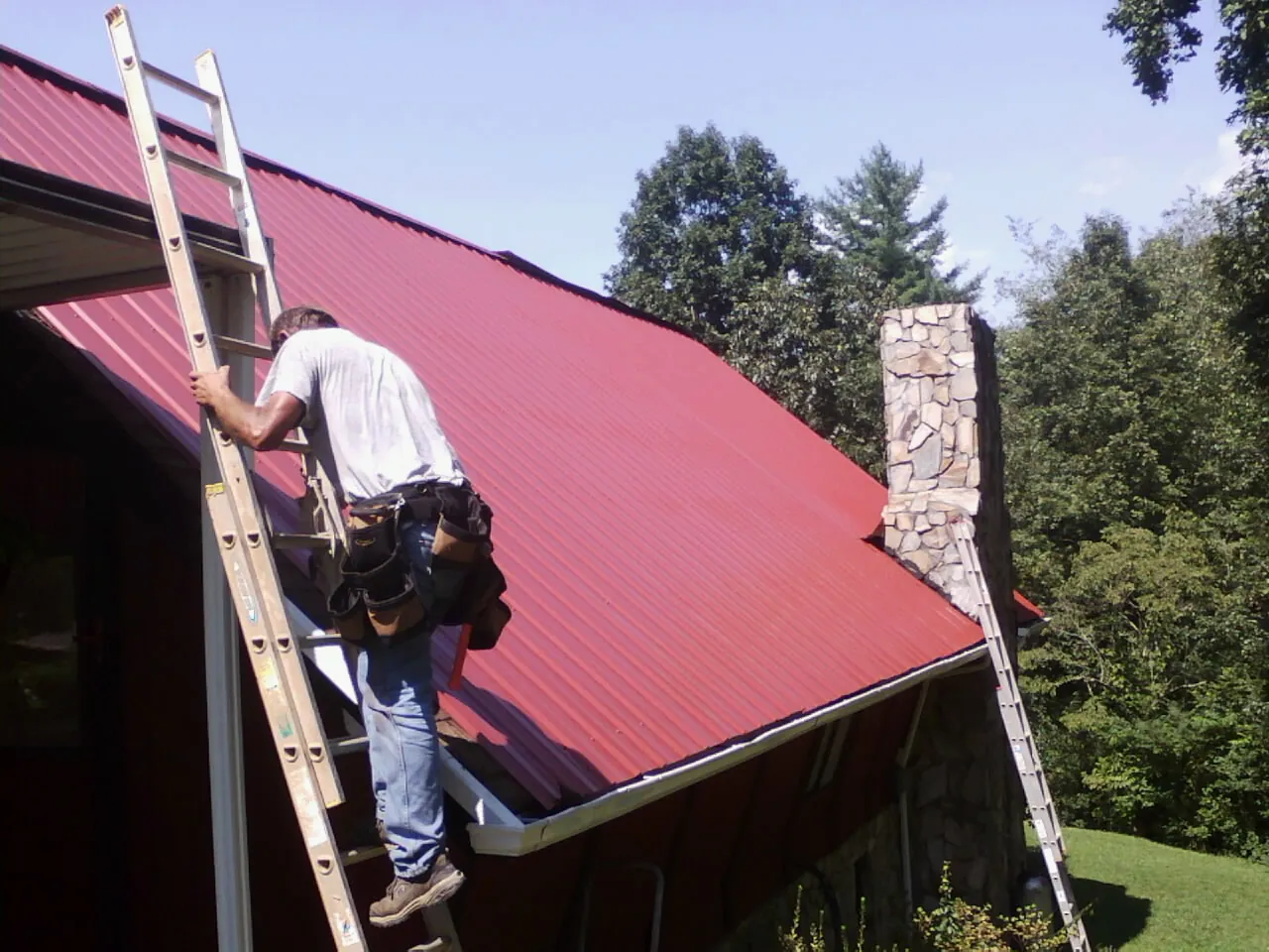 Roofer on a ladder beside a red metal roof and stone chimney — Soffit & Fascia Repair by Roofing Brothers in Corinth